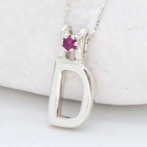 Op de afbeelding: Zilveren ketting met een hanger in de vorm van de letter D en een kleine rode edelsteen.