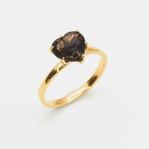 May include: A gold ring featuring a faceted heart-shaped smoky quartz gemstone set in a four-prong setting. The band is smooth and polished, showcasing a warm yellow gold tone that complements the dark brown, translucent hue of the central stone.