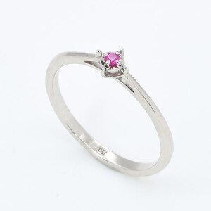 Puede incluir: Un anillo de banda de plata simple con una peque&ntilde;a piedra de rub&iacute; rosa redonda engastada en un engaste de cuatro garras.