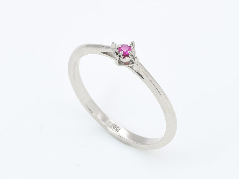 Puede incluir: Un anillo de banda de plata simple con una peque&ntilde;a piedra de rub&iacute; rosa redonda engastada en un engaste de cuatro garras.