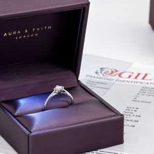 May include: A silver ring with a small diamond in the center, sitting in a purple velvet ring box. The box has the words "Aura & Faith London" on the inside of the lid. A diamond identification report is visible in the background.