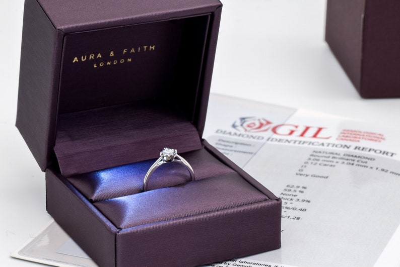 May include: A silver ring with a small diamond in the center, sitting in a purple velvet ring box. The box has the words "Aura & Faith London" on the inside of the lid. A diamond identification report is visible in the background.