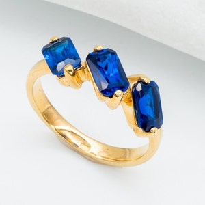 May include: A gold ring with three bright blue rectangular gemstones. The gemstones are arranged in a flowing design. The ring has a polished finish, reflecting light. The ring's design is eye-catching.