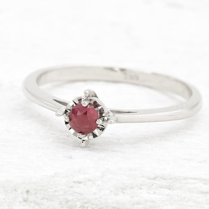 May include: A delicate silver ring with a red ruby gemstone set in a prong setting. The ring has a simple band and a small, round stone.