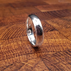 Könnte beinhalten: Ein polierter Silberring mit abgerundetem Band, der auf einer Holzoberfläche liegt. Der Ring reflektiert das Licht und zeigt seine glatte, glänzende Oberfläche. Der Ring scheint ein Ehering oder ein schlichtes, elegantes Schmuckstück zu sein.