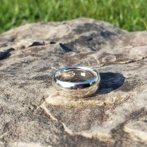 May include: A polished silver ring with a rounded design. The ring sits on a textured, grey stone surface, with green grass visible in the background. The ring's surface reflects the light, creating a bright appearance.