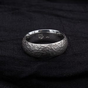 May include: A silver ring with a hammered texture. The ring is set against a black background. The ring's surface has a dimpled appearance, and the band is approximately 0.6 cm wide.