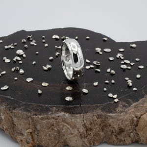 May include: A polished silver band ring with a smooth, rounded exterior surface. The ring is positioned upright, surrounded by numerous small, irregular silver-colored metal fragments scattered across the dark, glossy surface.