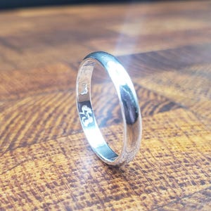 May include: A polished silver ring with a smooth, rounded band. The ring is set against a wooden surface with a natural grain pattern. The ring's surface reflects light, highlighting its shiny appearance.