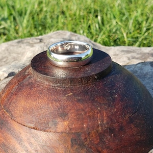 May include: A silver ring with a polished finish rests on a dark wooden object. The ring is circular and appears to be made of a shiny metal. The wooden object is a dark brown color and has a smooth, rounded shape. The background is blurred, with green grass visible.