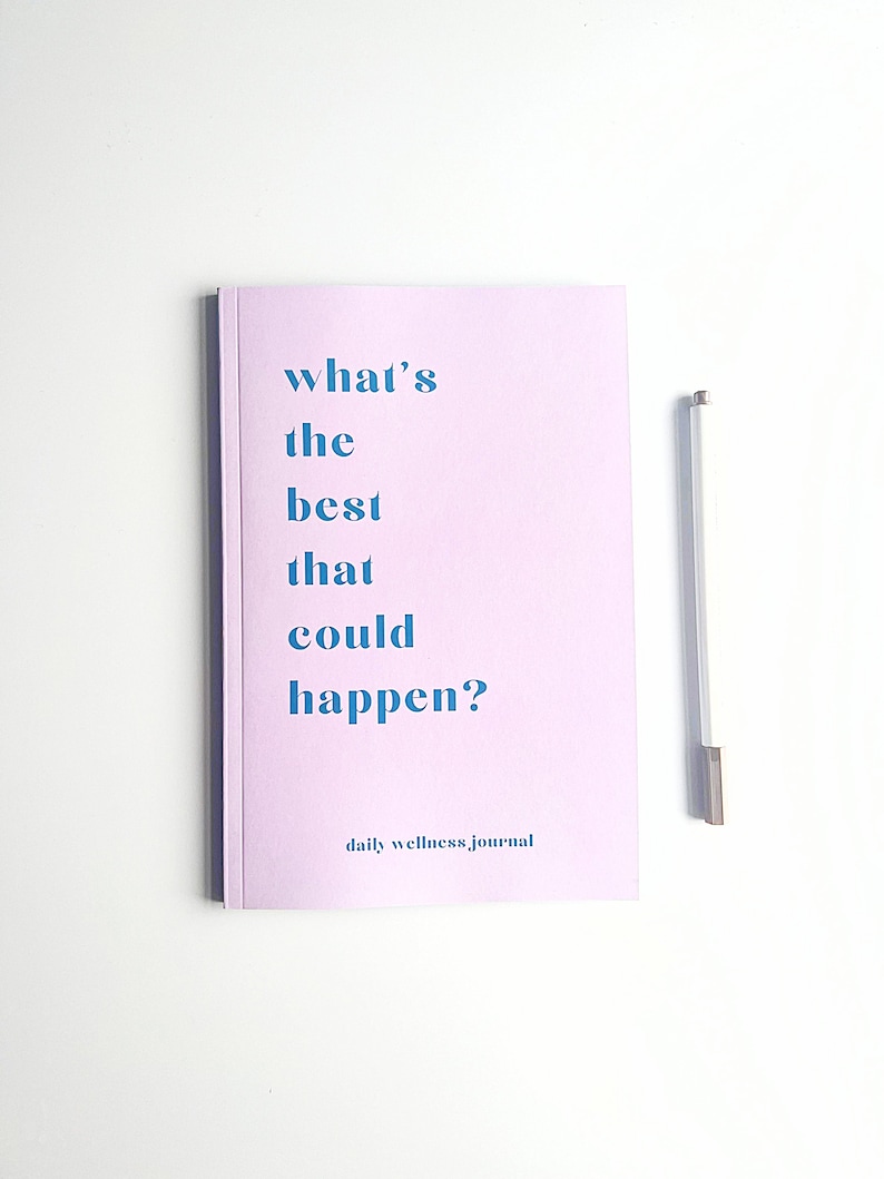 May include: A pink daily wellness journal with the text "what's the best that could happen?" in blue. The journal is on a white surface with a silver pen.
