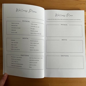 May include: A white wellness planner with a black and white design. The planner is divided into three sections: physical, mental, and emotional. Each section has a list of activities that can help improve well-being. The planner also includes a section for creating a personalized wellness plan.