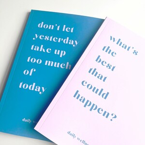 May include: Two pink and blue daily wellness journals with the text "don't let yesterday take up too much of today" on the blue journal and "what's the best that could happen?" on the pink journal.