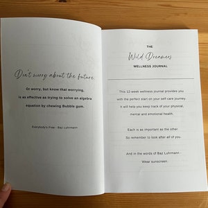 May include: An open book with a white background and black text. The left page has a quote about worrying, and the right page has the title "The Wild Dreamers Wellness Journal" and a description of the journal. The quote on the left page reads: "Don't worry about the future. Or worry, but know that worrying, is as effective as trying to solve an algebra equation by chewing Bubble gum. Everybody's Free - Baz Luhrmann"