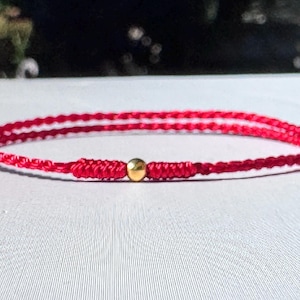 Puede incluir: Una pulsera fina de cordón rojo que presenta una pequeña cuenta esférica dorada y pulida en el centro. La pulsera está fabricada con hilo rojo trenzado con extremos anudados ajustables, creando un diseño circular simple y continuo.