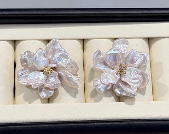 May include: A pair of earrings featuring iridescent, white baroque pearl petals arranged in a flower shape. Each earring has a small, textured gold-tone center. The earrings are displayed side-by-side on a cream-colored velvet jewelry insert.
