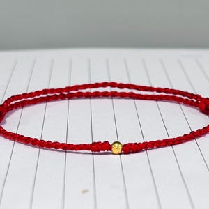 Puede incluir: Pulsera de hilo rojo con una pequeña cuenta dorada. La pulsera está hecha de dos hebras de cordón rojo, anudadas en cada extremo. La cuenta dorada está centrada en la pulsera.