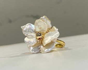 May include: A gold ring featuring a cluster of iridescent white baroque pearls arranged in a floral shape. The center of the pearl arrangement is accented with a small, smooth gold bead, and the pearls are secured to the gold band with thin gold wire.