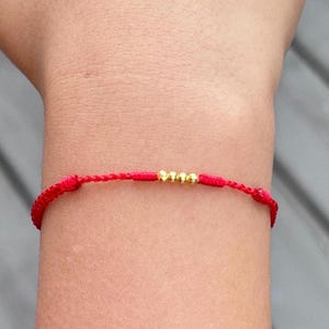 May include: A red cord bracelet featuring four small, round, gold-colored metallic beads centered on the string. The bracelet has a simple, adjustable design with knotted details on either side of the central gold beads.
