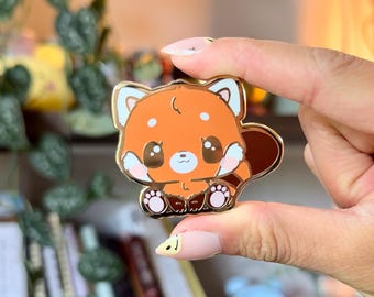 Könnte beinhalten: Eine goldfarbene Emaille-Anstecknadel mit einem Cartoon-Roten Panda mit braunem Fell, weißen Pfoten und großen braunen Augen. Der Rote Panda sitzt mit seinen Pfoten vor sich.