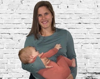 May include: A woman wearing a teal long-sleeve shirt and black jeans holds a baby in a rust-colored onesie. The baby is nursing. The background is a white brick wall.