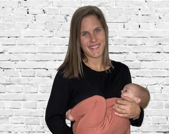 May include: A person wearing a black long-sleeve shirt and black jeans holds a baby wearing a rust-colored onesie. The background is a white brick wall.