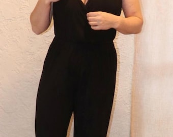 Könnte beinhalten: Schwarzer ärmelloser Jumpsuit mit V-Ausschnitt. Das einteilige Kleidungsstück hat ein Wickeloberteil und eine Hose mit geradem Bein. Die Person trägt braune Sandalen. Der Hintergrund ist eine strukturierte cremefarbene Wand und ein beiger Vorhang.