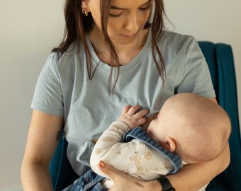 May include: A light blue t-shirt with a round neck. A baby wearing a patterned onesie and denim overalls is being held. The baby is nursing. The scene is set indoors.