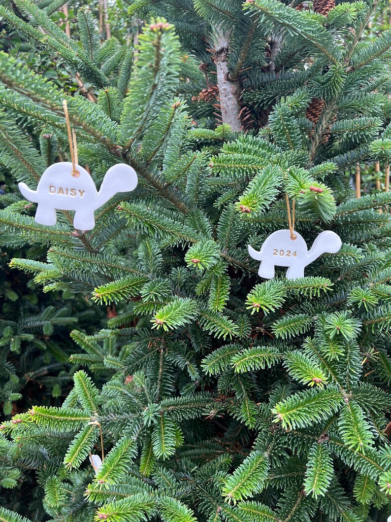 May include: Two white ceramic turtle ornaments hanging on a green Christmas tree. One ornament has the name "Daisy" and the other has the year "2024" in gold lettering. The ornaments are suspended by gold string.