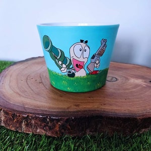 Puede incluir: Maceta de cerámica pintada a mano con un personaje de gusano de dibujos animados con una expresión de sorpresa, sosteniendo una pistola grande y un cañón. La maceta tiene un fondo azul claro con hierba verde en la parte inferior.