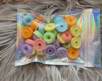 May include: A clear, iridescent bag filled with colorful, ring-shaped candies. The candies are in various pastel colors, including blue, orange, yellow, pink, green, and purple. The bag is sealed and sits on a fluffy, gray surface.
