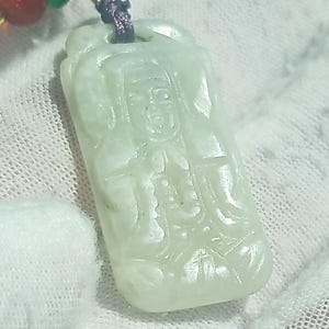 May include: A pale green jade pendant carved with a seated figure. The rectangular pendant is suspended from a purple cord with red and green beads. The carving details the figure's clothing and facial features. The pendant is likely of Asian origin.