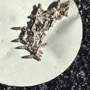 May include: A small, metallic silver-colored specimen with a dendritic, branching crystalline structure. The object features sharp, jagged points and a textured, reflective surface, appearing as a cluster of interconnected, needle-like metallic formations.