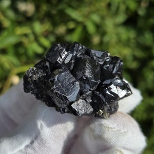 May include: A cluster of raw, dark black mineral crystals with a high-gloss, reflective surface. The specimen features sharp, angular geometric facets and a dense, compact structure, showcasing deep metallic tones and natural crystalline formations.