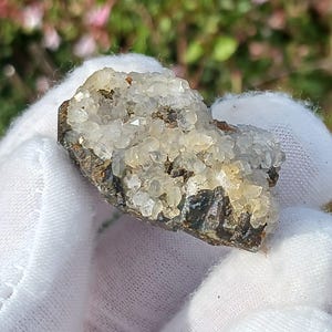 Peut inclure: Une petite roche grise foncée et rugueuse recouverte de cristaux de quartz clairs et brillants.