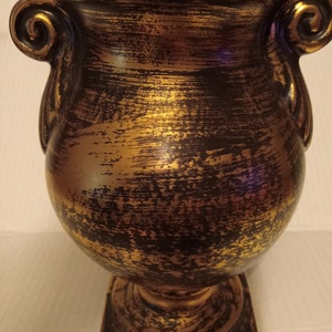 Puede incluir: Una urna decorativa con un acabado dorado y negro envejecido. La urna tiene un cuerpo redondeado, una base cuadrada y dos asas enrolladas. El acabado metálico le da a la urna una apariencia antigua. La urna es adecuada para la decoración del hogar.