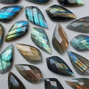 Puede incluir: Una colección de 15 cabujones de labradorita iridiscente en varios tonos de azul, verde y dorado. Las piedras están talladas en forma de lágrima y tienen un acabado liso y pulido.