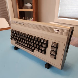 May include: A vintage Commodore 64 computer with a gray case and black keyboard. The keyboard has black keys with white lettering. The computer is resting on a black stand on a blue surface.