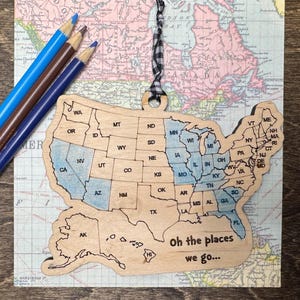 May include: Wooden ornament shaped like the United States of America with each state outlined. The ornament is painted in shades of blue and brown. The text "Oh the places we go..." is printed on the ornament.