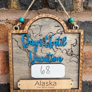 May include: A wooden wall hanging sign featuring a world map background, blue script text reading "Days Until Vacation," a white rectangular space with the number "68" written in gray, and a bottom plaque labeled "Alaska Our first cruise."