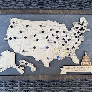 May include: Wooden United States Capitals travel tracker map with a dark wood frame. The map features state outlines, capital city names, and star-shaped pins. The text "UNITED STATES CAPITALS TRAVEL TRACKER" is displayed on a banner.