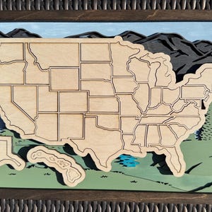 May include: A wooden map of the United States with state borders outlined in black. The map is set against a backdrop of green trees and mountains, with a blue lake in the lower right corner.