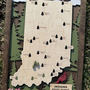 May include: Wooden Indiana State Parks map with laser-cut details. The map features a light wood state outline, dark trees, and park names. A red cardinal and pink flowers are at the bottom. The frame is dark brown.