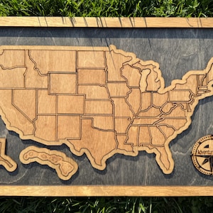 May include: A wooden map of the United States with each state outlined. The map is mounted in a dark wood frame with a compass in the bottom right corner that says "Adventure Awaits".