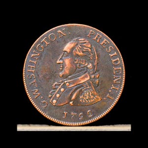 May include: A copper-colored circular coin featuring a profile portrait of George Washington facing left. The text G. WASHINGTON PRESIDENT I. is embossed along the upper edge, with the date 1792 inscribed at the bottom of the coin face.