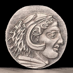 May include: A silver coin with a detailed profile of a person wearing a helmet and a crown. The person's face is turned to the side, and their hair is flowing.