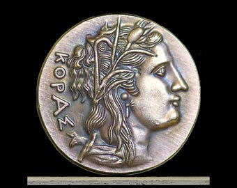 May include: A circular bronze-toned medallion featuring a relief profile of a figure facing right, adorned with a leafy headpiece. The Greek text KOPΑΣ is embossed vertically along the left edge. The surface has a metallic, textured finish.