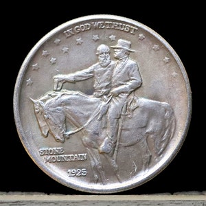 May include: A silver coin with a portrait of two men on horseback. The coin reads "In God We Trust" and "Stone Mountain 1925".