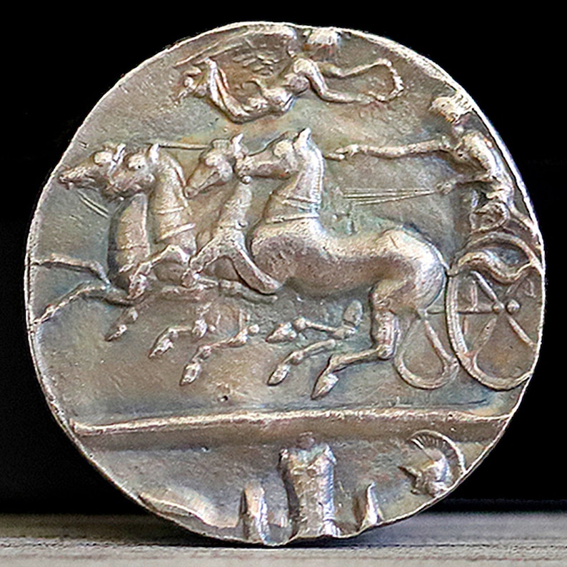 Puede incluir: Moneda de plata con la representaci&oacute;n de un carro tirado por cuatro caballos, con una figura alada arriba. El conductor del carro es visible en el lado derecho de la moneda.