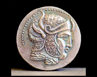 May include: A circular bronze-toned medallion featuring a relief profile of a figure wearing a textured helmet with curved horn-like elements. The design includes a dotted border along the upper edge and a metallic finish with dark patina.
